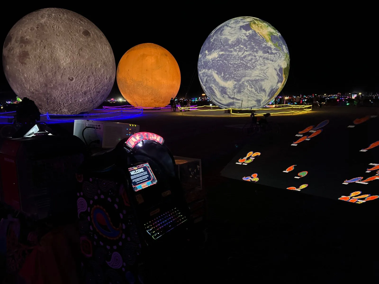 An AFTM on the playa at night, giant moon, sun and earth sculptures behind, paisleys projected on the ground
